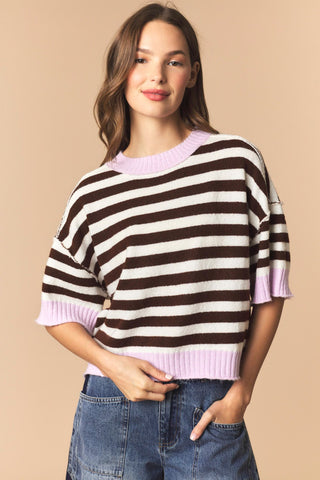 A woman wearing a brown and white horizontally striped knit sweater with light purple ribbed trim, paired with blue jeans, against a solid beige background.