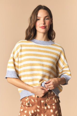 A woman wearing a yellow and white striped knit sweater with light blue ribbed trim, paired with brown patterned pants, against a solid beige background.