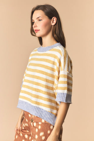 A woman wearing a yellow and white striped knit sweater with light blue ribbed trim, paired with brown patterned pants, against a solid beige background.
