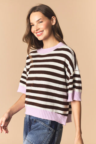 A woman wearing a short-sleeve knit sweater with horizontal dark brown and off-white stripes, finished with light purple ribbed trim at the crew neck, sleeve cuffs, and hem, paired with blue jeans against a solid beige background.