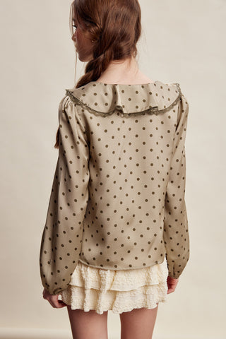 A woman shown from the back wearing a beige long-sleeve blouse with dark brown polka dots and a large ruffled collar, paired with a cream ruffled skirt, against a plain beige studio background.