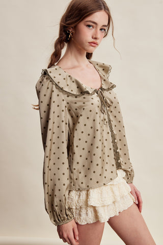A woman wearing a beige long-sleeve blouse with brown polka dots and a large ruffled collar, paired with a cream ruffled skirt, against a plain beige studio background.