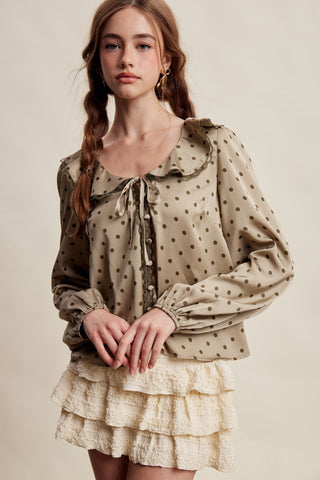A woman modeling a beige long-sleeve blouse with dark brown polka dots and a ruffled tie neckline, paired with a cream ruffled mini skirt, against a plain beige studio background.