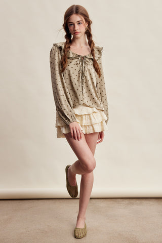 A woman modeling a beige long-sleeve blouse with black polka dots, a ruffled tie neckline, and a matching ruffled mini skirt, paired with beige woven flat shoes against a neutral studio backdrop.