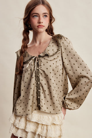 A woman wearing a beige blouse with dark brown polka dots, featuring a ruffled collar, tie detail at the neckline, and long voluminous sleeves, paired with a cream ruffled skirt against a warm beige studio background.