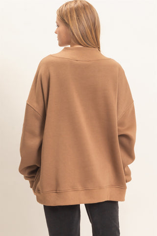 A woman shown from the back wearing an oversized tan crewneck sweatshirt and dark pants, photographed against a plain light background.