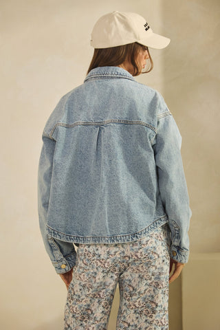 Person wearing a denim jacket and floral pants with a beige cap on a plain background