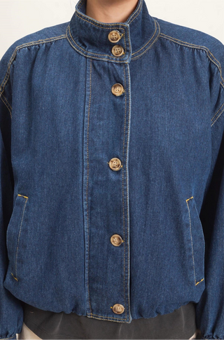 A cropped blue denim jacket with a high stand collar, front button closure, and side pockets, shown on a model against a plain light background.