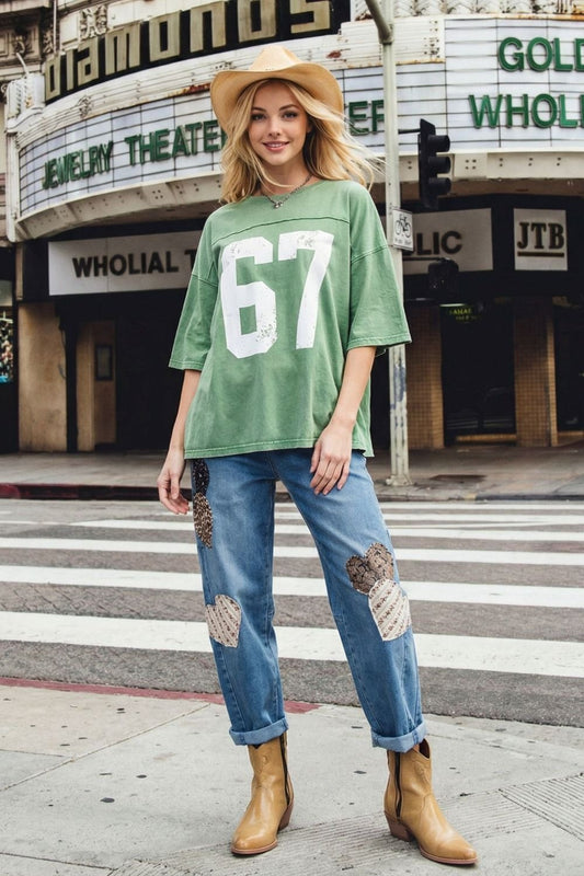 Woman wearing a green oversized shirt with the number 67, blue jeans with patches, and tan boots on a city street.