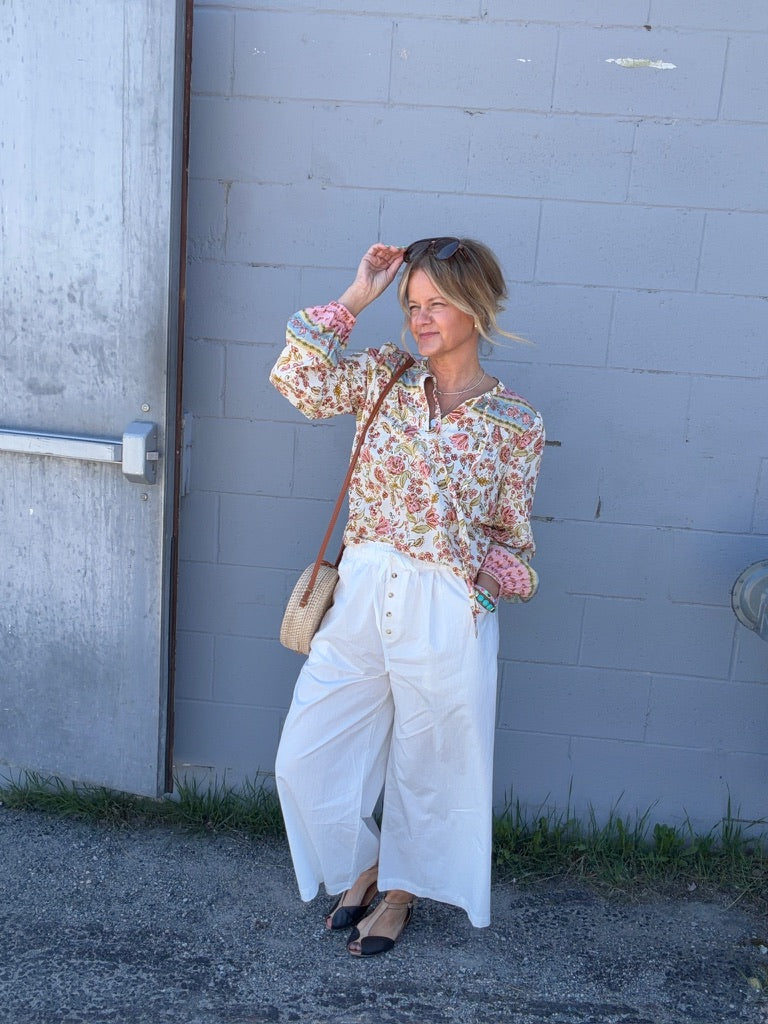 Women wearing a floral print blouse and white pants on a grey background.