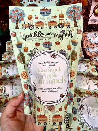 A hand holding a bag of Pickle and Myrh Caramel Apple Caramels. The packaging is a light mint green with autumn-themed illustrations of pumpkins, trees, and trains, and is held against a background of more packaged caramels.