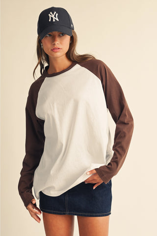 A woman wearing a two-tone raglan long-sleeve shirt with a white body and brown sleeves, paired with a navy baseball cap featuring a white New York Yankees logo and a dark denim mini skirt, set against a solid beige background.