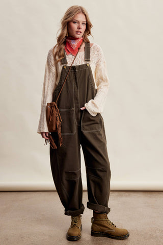 A woman modeling a pair of dark brown utility overalls with a cream knit sweater, a red bandana, and brown boots against a neutral studio backdrop.