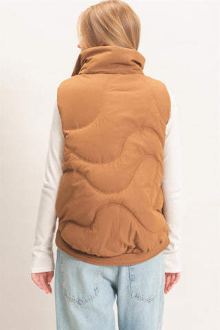 A woman viewed from behind wearing a camel brown quilted puffer vest over a white long-sleeve top and light blue jeans, against a plain white studio background.