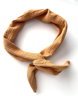 A textured, gauze-like fabric headband or scarf in a warm camel tan color, styled in a loop with a knot at the bottom, set against a plain white background.
