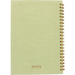 A light sage green linen-covered spiral notebook with a gold wire binding and the brand name 'danica' printed near the bottom.