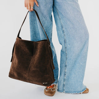 A person wearing light blue wide-leg jeans and brown shoes carries a large dark brown suede shoulder bag against a plain white background.