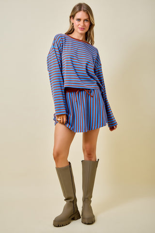 A woman modeling a blue and orange striped long-sleeve top and matching striped mini skirt with a drawstring, paired with olive green knee-high boots, against a solid beige background.