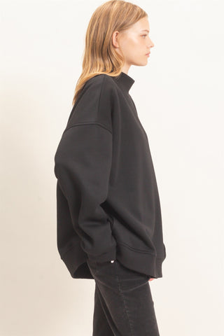 A woman shown in side profile wearing an oversized black sweatshirt and black jeans against a plain off-white studio background.