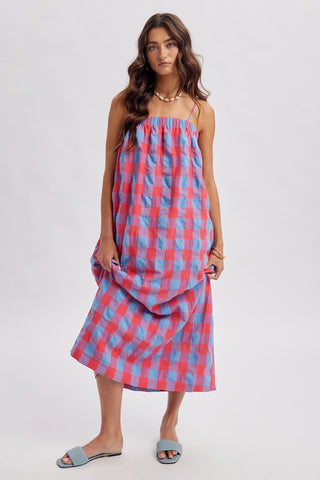 A woman modeling a sleeveless midi dress with a pink and blue gingham pattern, paired with gray slide sandals, against a plain light gray studio background.