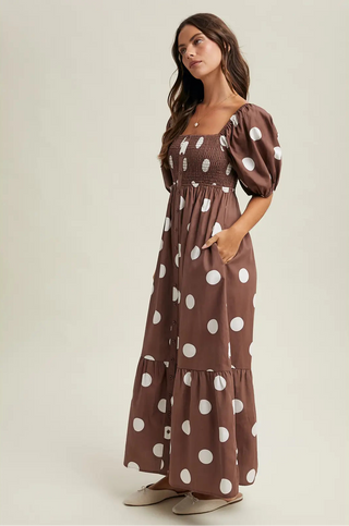 A woman modeling a brown smocked maxi dress with large white polka dots, standing against a plain light beige studio background.
