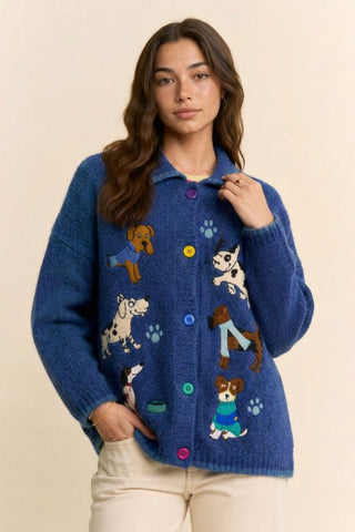A woman wearing a blue fuzzy knit cardigan with colorful embroidered dog illustrations and multicolored buttons, paired with cream pants, against a plain beige background.
