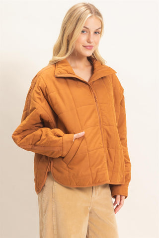A woman wearing a cropped quilted jacket in a warm camel-brown color, paired with beige pants, photographed against a plain off-white studio background.