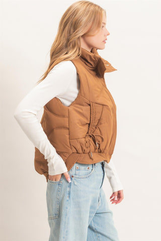A woman shown in side profile wearing a brown puffer vest over a white long-sleeve top and light blue jeans, photographed against a plain light background.