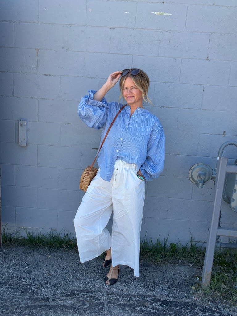 Woman in a blue shirt and white pants standing against a light blue brick wall.
