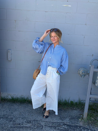 Woman in a blue shirt and white pants standing against a light blue brick wall.