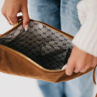 A close-up of a person holding open a brown suede pouch or clutch, showing a black patterned interior lining with a gold zipper. The person is wearing blue jeans and a white sweater.