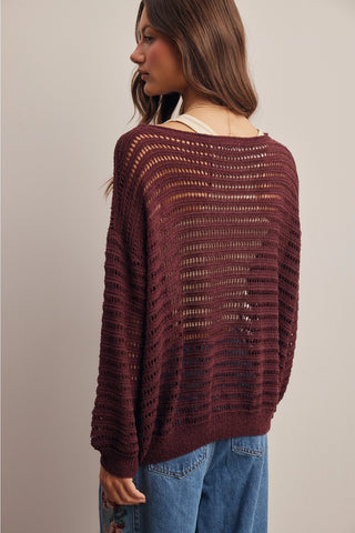 A woman shown from the back wearing a loose, open-knit maroon sweater over a white undershirt and blue jeans, set against a plain light gray studio background.