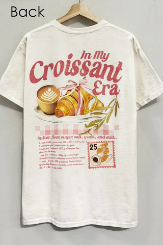 An off-white short-sleeve graphic t-shirt hanging on a wooden hanger against a neutral gray wall. The shirt features a vintage-style print with the text 'In My Croissant Era' in red, accompanied by illustrations of a croissant, coffee, and a recipe list.