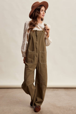 A woman wearing olive green wide-leg canvas overalls, a cream long-sleeve blouse, a brown wide-brim hat, and brown shoes, standing against a neutral beige studio backdrop.