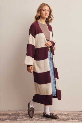 A woman models a long, open-front knit cardigan with wide horizontal stripes in deep burgundy and cream. She is styled with a taupe top, light-wash blue jeans, white socks, and dark brown loafers against a neutral studio backdrop.