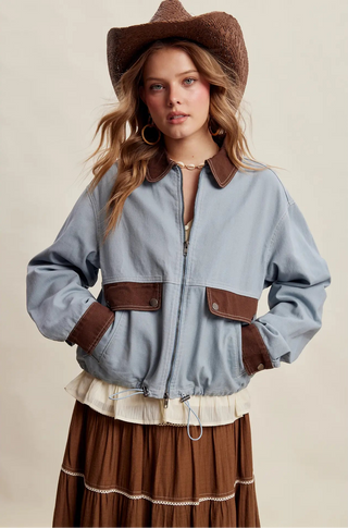 A woman wearing a light blue denim jacket with brown faux-leather trim, a brown cowboy hat, and a brown pleated skirt, posed against a solid beige studio background.