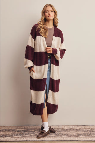 A woman models a long, open-front knit cardigan with wide horizontal stripes in deep burgundy and cream. She is wearing a taupe top, blue jeans, white socks, and brown loafers, standing against a neutral beige wall.