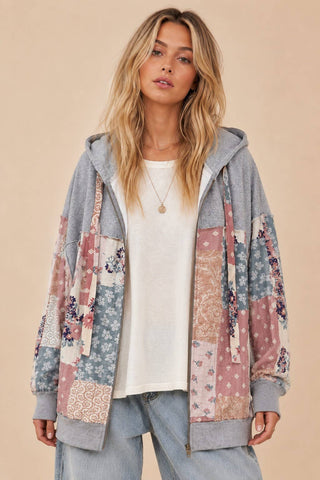 A woman wearing a gray zip-up hoodie with a colorful patchwork floral front panel, styled over a white t-shirt and light blue jeans against a solid beige background.