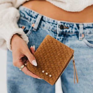 A person holding a small brown woven zip wallet with three rhinestone studs, wearing blue jeans and a white fuzzy sweater.