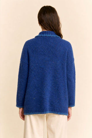 A person shown from the back wearing a textured royal blue knit cardigan sweater and cream-colored pants, set against a plain beige background.