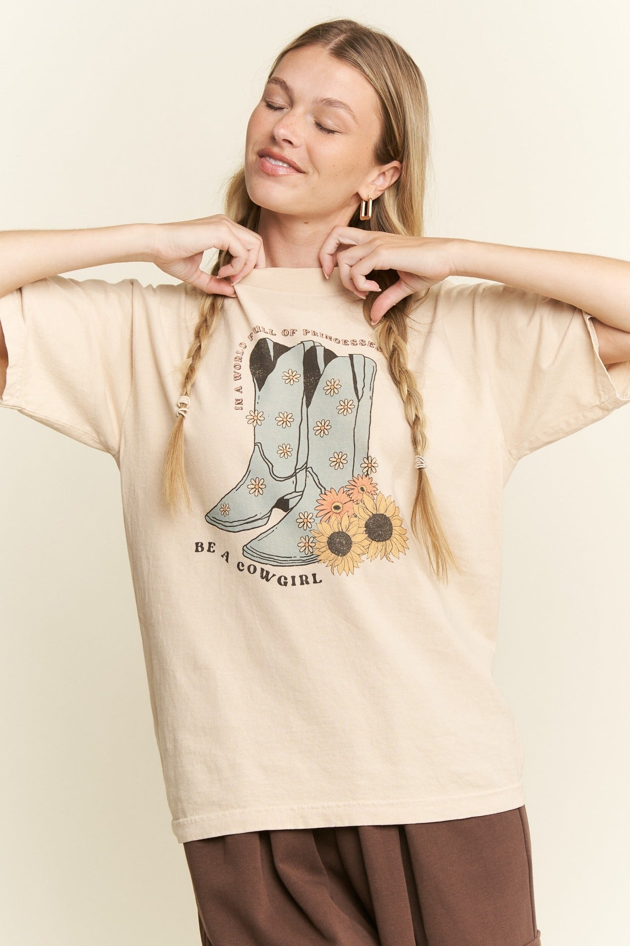Woman wearing a beige t-shirt with cowboy boot and sunflower design, text 'Be a Cowgirl', on a beige background.
