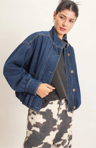A woman modeling a cropped dark blue denim jacket over a black top and a black-and-white cow-print skirt, photographed against a plain light background.