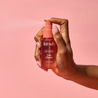 Hand holding a 'kitsch' Tart Cherry hair perfume bottle against a pink background