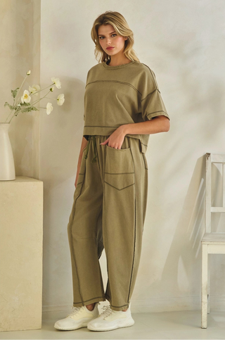 A woman models a matching two-piece lounge set in a warm khaki/olive green color, featuring a cropped short-sleeve top and wide-leg pants with side pockets, styled with white sneakers in a minimalist indoor setting.