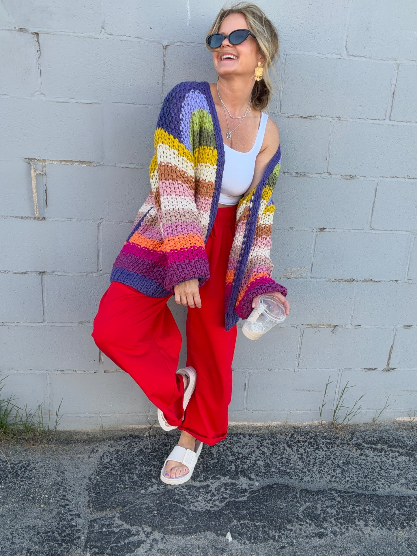 Woman wearing a colorful striped cardigan and red pants against a gray wall.