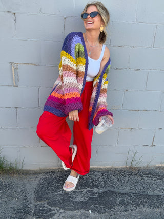 Woman wearing a colorful striped cardigan and red pants against a gray wall.