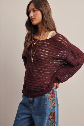 A woman wearing a loose, open-knit burgundy sweater with a ribbed texture, styled with blue jeans featuring floral embroidery on the leg and gold necklaces, set against a plain beige studio background.
