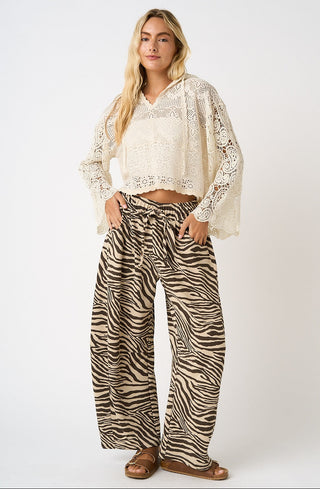 A woman modeling a bohemian-style outfit consisting of a cream lace long-sleeve crop top and high-waisted brown and beige zebra-print wide-leg pants, paired with brown leather sandals, against a light gray studio background.