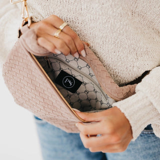 A close-up of a person holding open a blush pink woven crossbody bag, showing the interior lining and a black brand patch with a white monogram logo. The person is wearing a cream knit sweater and blue jeans.