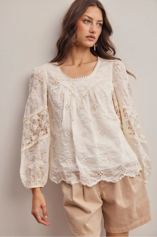 A woman wearing a cream-colored eyelet lace blouse with long sleeves and beige shorts, standing against a neutral beige background.